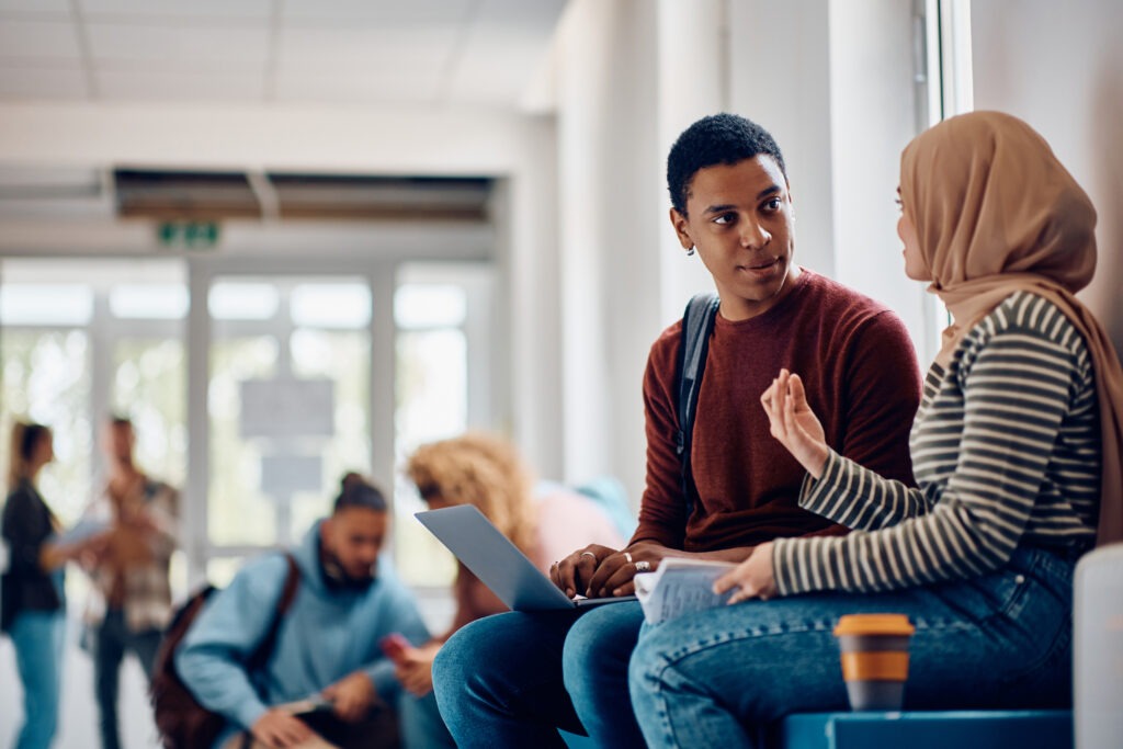 Black student using laptop while talking to female friend at university hallway.