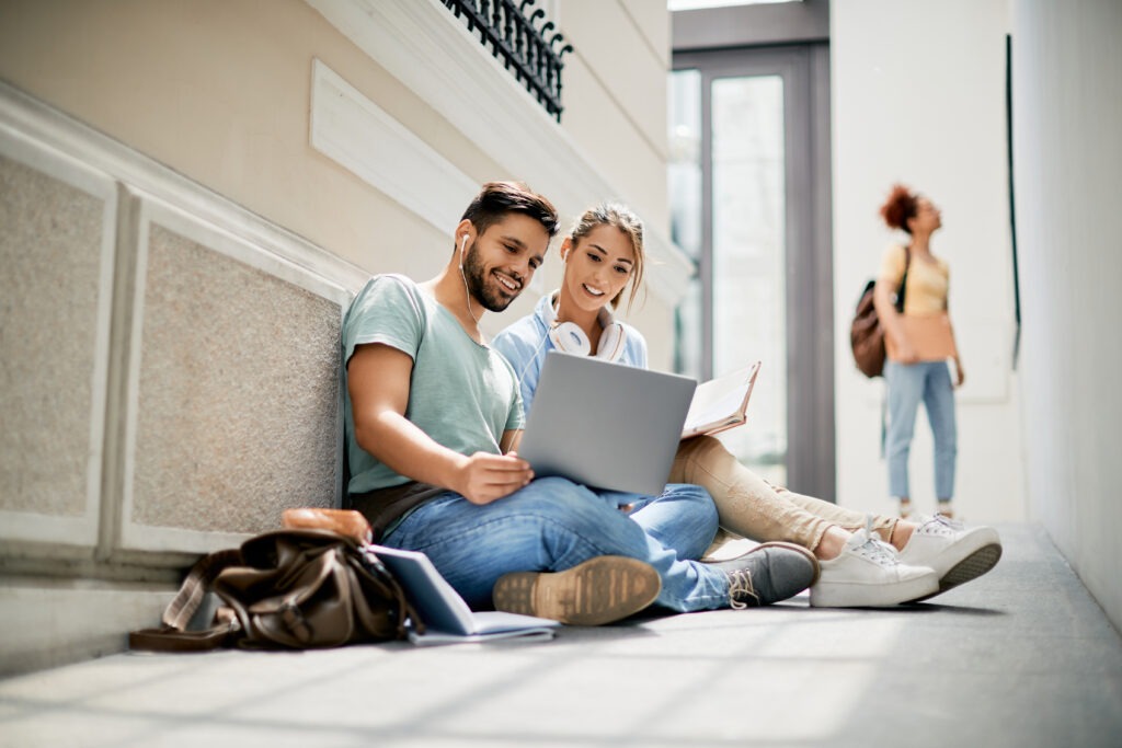 Happy university friends using laptop while studying in a hallway.