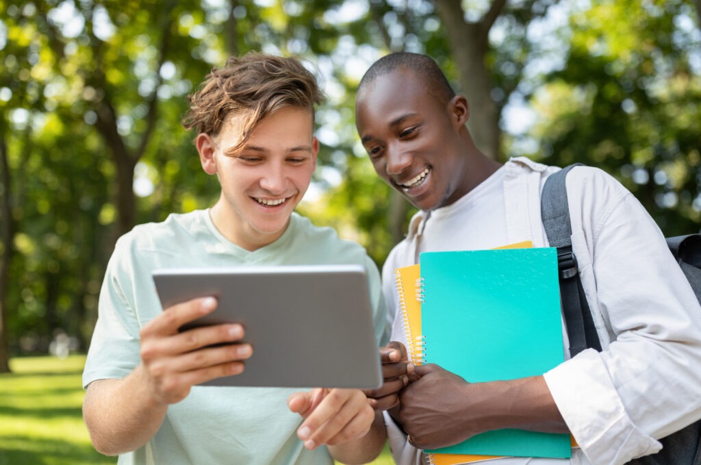 Student guy showing his project on digital tablet to friend, walking outdoors in college campus