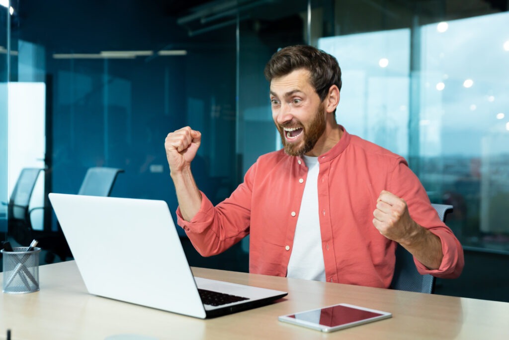 A young man sits in the office, looks at the laptop monitor and rejoices. Makes victorious hand