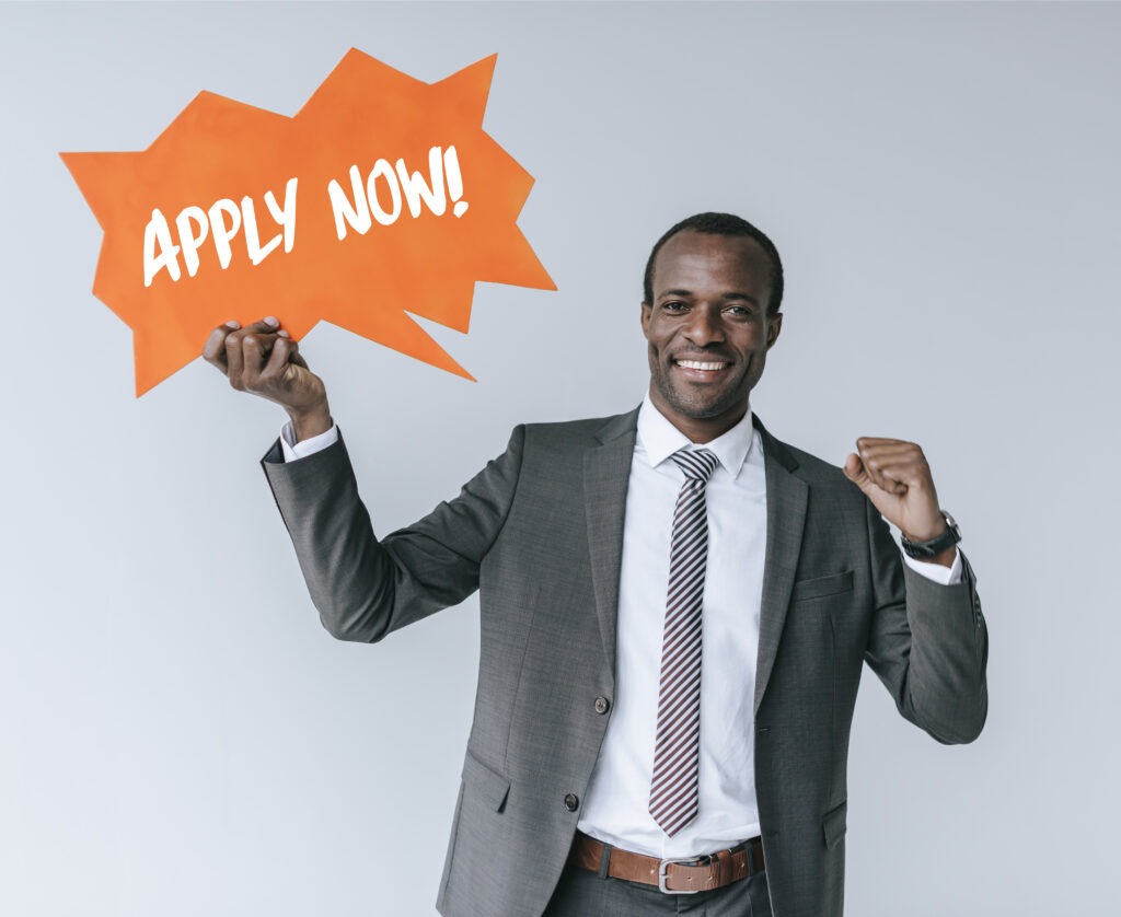 portrait of smiling african american businessman with apply now card isolated on grey
