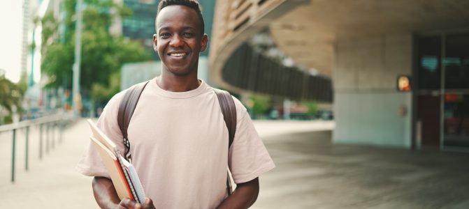 Young african student stands outside of university, looks at the camera, smiles