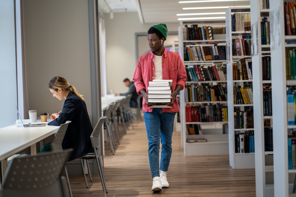 African American international student guy walking with stacked books through college library