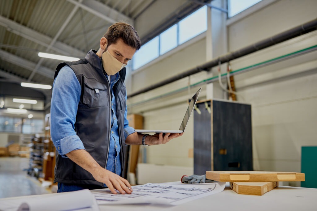 Carpenter with face mask examining blueprints while using computer at woodworking facility.