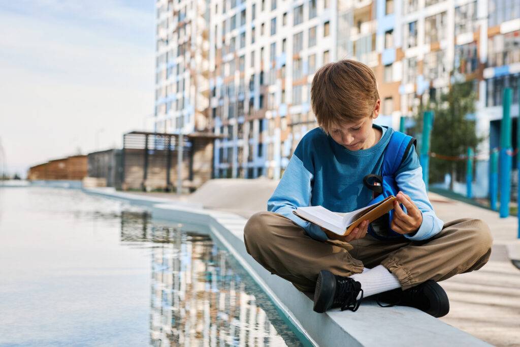 Cute diligent schoolboy in casualwear sitting by riverside and reading